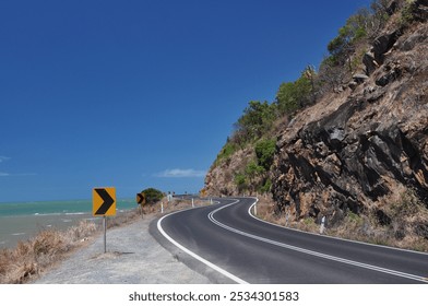 A scenic coastal road with a smooth, winding asphalt stretch running alongside steep rocky cliffs and overlooking the turquoise waters of the Coral Sea, under a vibrant blue sky. - Powered by Shutterstock