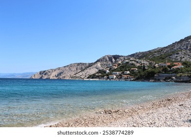 A scenic coastal view with clear turquoise waters, a rocky shoreline, and a small village nestled against rugged hills under a bright blue sky - Powered by Shutterstock