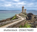 A scenic coastal view featuring a lighthouse Le Petit Minou in Plouzane on a rocky promontory surrounded by lush greenery and wildflowers, with the ocean and a cloudy sky in the background.