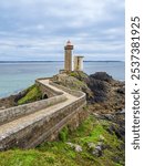 A scenic coastal view featuring a lighthouse Le Petit Minou in Plouzane on a rocky promontory surrounded by lush greenery and wildflowers, with the ocean and a cloudy sky in the background.