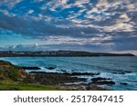 A scenic coastal view featuring rocky shores, gentle waves, and a distant town under a cloudy sky. At Little Fistral Beach in Newquay, Cornwall, UK