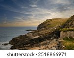 A scenic coastal view featuring rocky cliffs, a sandy beach, and a calm sea under a cloudy sky. At Tintagel, Cornwall, UK.