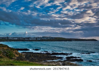 A scenic coastal view featuring rocky shores, gentle waves, and a distant town under a cloudy sky. At Little Fistral Beach in Newquay, Cornwall, UK - Powered by Shutterstock