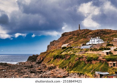 A scenic coastal view featuring a rocky shoreline, a white house, and a historic mine chimney on a hill under a dramatic sky with clouds.At Cape Cornwall, UK. - Powered by Shutterstock