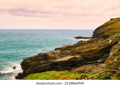 A scenic coastal view featuring rocky cliffs, turquoise waters, and a cloudy sky. At Tintagel, Cornwall, UK. - Powered by Shutterstock