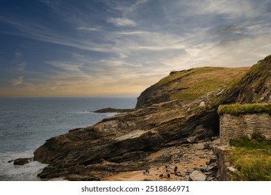 A scenic coastal view featuring rocky cliffs, a sandy beach, and a calm sea under a cloudy sky. At Tintagel, Cornwall, UK. - Powered by Shutterstock