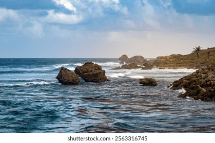 A scenic coastal view featuring rocky formations, gentle waves, and a lush green hillside under a cloudy sky. The ocean reflects various shades of blue and green. - Powered by Shutterstock