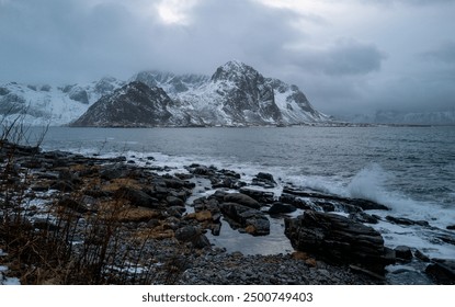 A scenic coastal view featuring a snow-capped mountain, rocky beach, stormy sky, and dynamic ocean, with no visible text in the image, Lofoten, Norway - Powered by Shutterstock