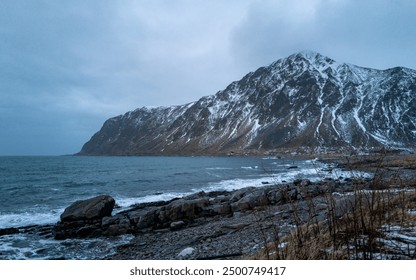 A scenic coastal view featuring a snow-capped mountain, rocky beach, stormy sky, and dynamic ocean, with no visible text in the image, Lofoten, Norway - Powered by Shutterstock