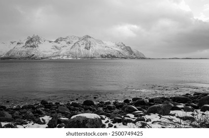 A scenic coastal view featuring a snow-capped mountain, rocky beach, stormy sky, and dynamic ocean, with no visible text in the image, Lofoten, Norway - Powered by Shutterstock