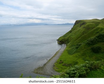 A scenic coastal view on the Isle of Skye, with lush green cliffs descending to a pebble beach and calm sea, under a cloudy sky with distant mountains visible on the horizon. - Powered by Shutterstock