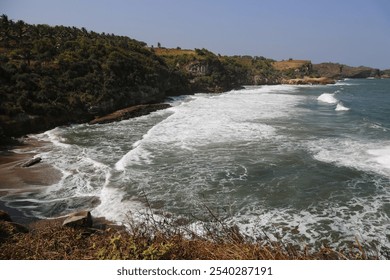 A scenic coastal view with a rocky shoreline, crashing waves, and lush green vegetation in the background. The sky is clear and blue, creating a beautiful contrast with the turquoise ocean water. - Powered by Shutterstock