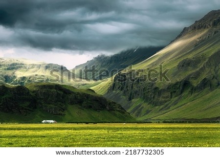 Scenic of dramatic Icelandic mountain with sunlight shining through storm clouds and house on field in summer at Iceland