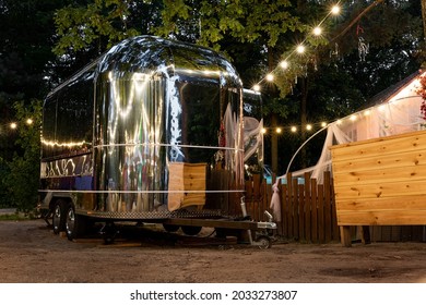 Scenic evening view of vintage futuristic silver metal camper van trailer parked near wooden fence at countryside farm or ranch against light garland forest tree. Retro style fodtruck caravan vehicle