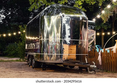 Scenic evening view of vintage futuristic silver metal camper van trailer parked near wooden fence at countryside farm or ranch against light garland forest tree. Retro style fodtruck caravan vehicle