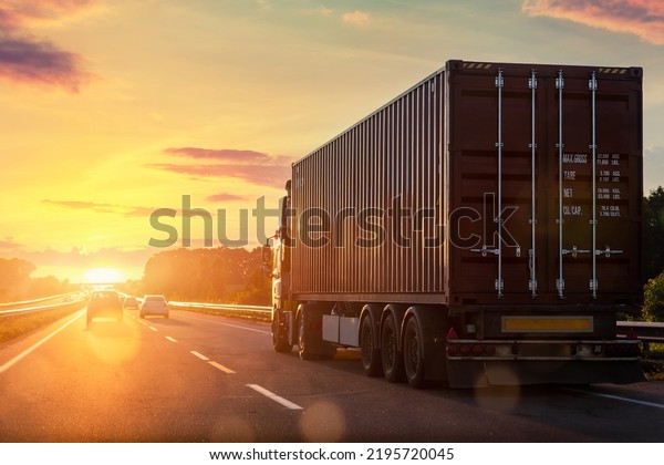 Scenic front view big long heavy semi-treailer\
truck with sea shipping container driving highway dramatic warm\
morning evening sunrise sun sky sunset. Cargo transport industry\
background concept
