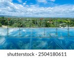 A scenic glass skywalk with reflective surface offers panoramic views of lush green landscapes and distant mountains under a bright blue sky.