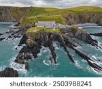 Scenic Hartland Quay with hotel on the cliff. Aerial view from the sea on rock formations and colorful hilly seaside landscape with green grass in North Devon UK. Cloudy day in summer. Cars parked. 