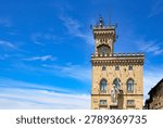 A scenic horizontal low-angle view showcasing the historic Palazzo Pubblico, the town hall and official government building of San Marino, located in Piazza della Liberta. The image highlights the stone neo-gothic facade, crenellated clock tower, and the prominent white Carrara marble Statue of Liberty (Statua della Libertà) standing proudly in front