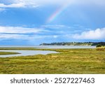 A scenic landscape in Brittany featuring a grassy marshland with a distant rainbow arching over a calm body of water, surrounded by lush greenery and a partly cloudy sky.