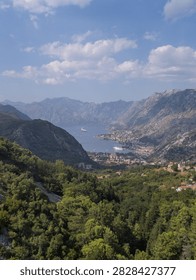 Scenic landscape featuring cliffs, forests, a coastal town, and a calm sea in a protected bay, distant mountains and a clear azure sky over blue water
