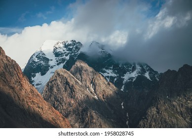 Scenic landscape with great rocks and snowy mountains in sunlight in low clouds. Wonderful view to mountain top with snow in sunshine in cloudy sky. Awesome scenery with sunlit snow-white pinnacle.