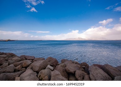 Scenic Landscape Shot Of Calm Ocean Waters Shot From A Shore With Big Rocks In Foreground. The Blue Skies & White Clouds Are Visible Till Horizon With An Island At Far End. Peaceful & Tranquil Shot.