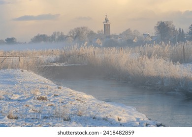 scenic misty and snowy sunrise over the church St. Marien and residential houses in Brake Unterweser (Germany) - pastures and the small river Rönnel can be seen in foreground