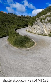 Scenic mountain road with a sharp switchback turn leading to Sokraki village in Corfu, Greece. Winding asphalt path surrounded by lush green vegetation, rocky hills, and a blue sky