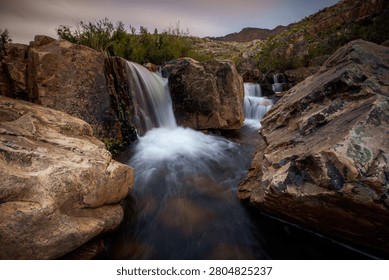 A scenic mountain stream flows over layered rock formations creating a series of small cascading waterfalls, surrounded by boulders and sparse vegetation in an arid rocky landscape.