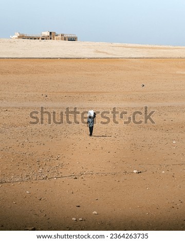 Scenic panorama of an Egyptian man walking in the middle of the sandy desert. He is wearing typical Egyptian clothing, with tunic and sandals. He is carrying a jerrycan with water on his back.