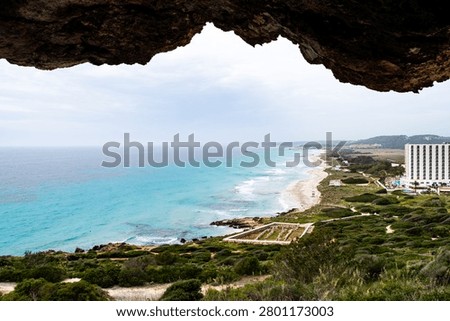 Scenic panoramic view of Son Bou beach and ancient ruins in Menorca, framed by the dark interior of a stone cave