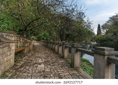 Scenic pathway features cobblestone surface bordered by stone walls, surrounded by vibrant greenery and trees, leading to a serene water body, creating a peaceful and inviting atmosphere