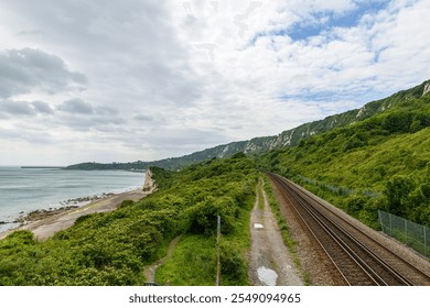 A scenic railway line running parallel to the coastline, bordered by lush greenery and dramatic white cliffs, with views of the English Channel under a bright sky - Powered by Shutterstock