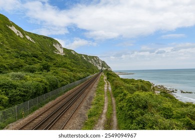 A scenic railway line running parallel to the coastline, bordered by lush greenery and dramatic white cliffs, with views of the English Channel under a bright sky - Powered by Shutterstock