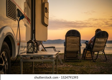 Scenic RV Campsite Pitch. Camper Van In the Recreational Vehicles Park. Woman Relaxing on a Chair and Watching Sunset Over the Sea.
