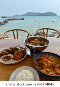 Scenic seaside dining experience with authentic Thai cuisine served on a wooden table overlooking a calm turquoise ocean and distant island landscape. The composition features a variety of traditional dishes including seafood tom yum soup, stir-fried shrimp in rich sauce, Thai fish cakes with dipping sauce, and a bowl of steamed jasmine rice. Framed by minimalist chairs and a panoramic coastal view, this image captures the essence of tropical lifestyle, beachside restaurant culture, and relaxed vacation dining in Thailand. Perfect for travel, food, hospitality, and lifestyle concepts, highlighting fresh ingredients, local flavors, and oceanfront ambiance.