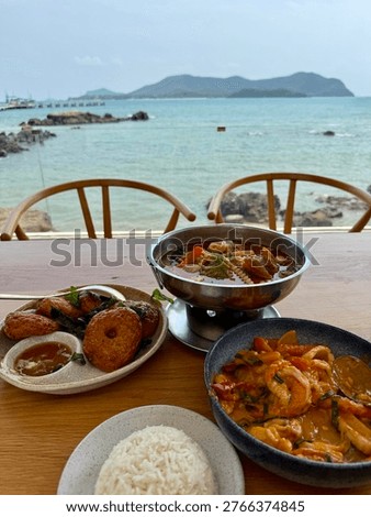 Scenic seaside dining experience with authentic Thai cuisine served on a wooden table overlooking a calm turquoise ocean and distant island landscape. The composition features a variety of traditional dishes including seafood tom yum soup, stir-fried shrimp in rich sauce, Thai fish cakes with dipping sauce, and a bowl of steamed jasmine rice. Framed by minimalist chairs and a panoramic coastal view, this image captures the essence of tropical lifestyle, beachside restaurant culture, and relaxed vacation dining in Thailand. Perfect for travel, food, hospitality, and lifestyle concepts, highlighting fresh ingredients, local flavors, and oceanfront ambiance.
