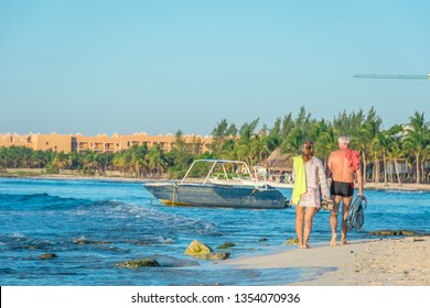 A Scenic Shot Of An Older Couple Walking The White Sand Shoreline Of A Beach In Mexico, With Fishing Boats Floating On The Turquoise Waters Of The Caribbean, And Palm Trees In The Background.  