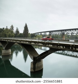 A scenic shot of a red vehicle crossing a modern concrete bridge over a calm river, with a historic railway bridge and lush greenery in the background - Powered by Shutterstock