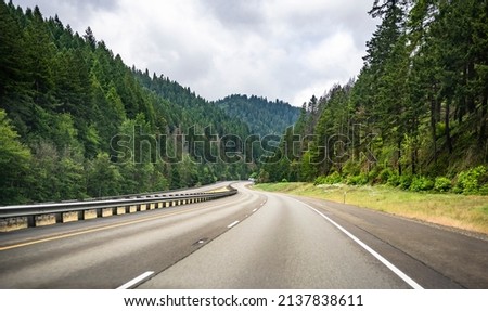 Scenic summer landscape with a disappearing around the corner winding mountain highway road with markings and a security divided fence and green forest trees on the slopes of mountain in Oregon