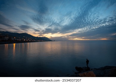 Scenic Sunset Landscape over Black Sea Coast with Silhouette of Fisherman on Rocky Shore, Dramatic Clouds and City Lights of Rize, Turkey - Powered by Shutterstock