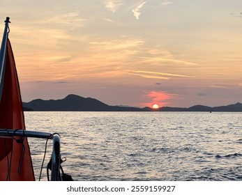 Scenic sunset over calm sea with mountains in the distance. Silhouette of a boat railing and red flag in the foreground. Vibrant sky with orange and pink hues reflects on the water surface. - Powered by Shutterstock