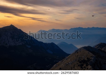 Scenic sunset over a dramatic mountain range with layered ridges fading into the distance. Soft, warm light bathes the sky in golden and purple hues, while birds soar peacefully above the tranquil lan