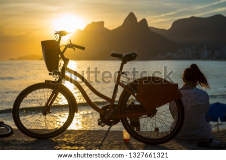 Scenic sunset view of Ipanema Beach with Two Brothers Mountain towering above the silhouette of a woman with a bicycle sitting at the Arpoador overlook in Rio de Janeiro, Brazil