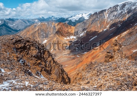 Scenic top view from stony precipice edge to multicolor big rocky ridge in freshly fallen snow and giant snow-capped mountain peak in low clouds. Snow-covered multi-color sharp rocks in huge mountains