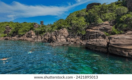 A scenic tropical cove with people snorkeling in clear blue water near rugged rocks under a bright blue sky, ideal for summer vacation concepts
