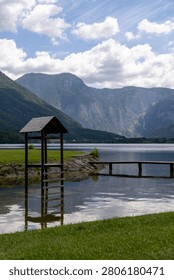 A scenic vertical shot of a small rustic wooden tower with a roof standing in the clear shallow water of Lake Hallstatt in Austria. In the background, a simple wooden pier extends into the calm lake surface, bordered by a stone-reinforced grassy shore. Majestic alpine mountains covered with green forests and rocky details rise high under a blue sky filled with bright white clouds. The peaceful water beautifully reflects the wooden structure and the clouds, capturing a tranquil summer day in the Salzkammergut region, perfect for European travel, tourism, and nature vacation concepts