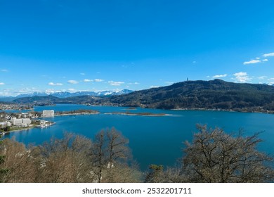 Scenic view of alpine Lake Wörth surrounded by snow-capped mountains Karawanks in Carinthia, Austria, Europe. Viewing tower Pyramidenkogel reflected in calm water surface. Hohe Gloriette, Pörtschach - Powered by Shutterstock