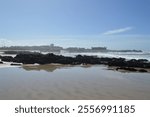 A scenic view of a beach in Porto, Portugal, with dark rocky outcrops and shallow water reflecting the clear blue sky. A historic fortress and distant buildings complete the coastal landscape